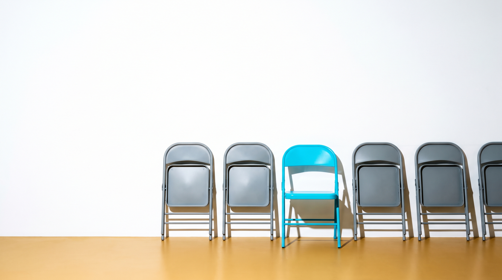 Seven folding chairs against a white wall, six grey and facing away, one cyan facing the camera
